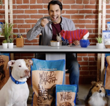 Man eating pet food behind two dogs and many dog food bags