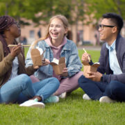 Multiethnic students talking and eating takeaway meal relaxing in summer park