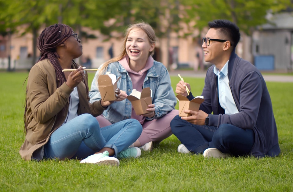 Multiethnic students talking and eating takeaway meal relaxing in summer park